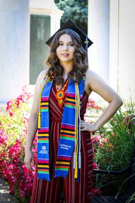 A person wearing a graduation cap and vibrant stole stands confidently amidst colorful flowers and greenery. The stole reads 'Excellence in Diversity' and is decorated with bright, multicolored stripes. The person is dressed in a striped, sleeveless outfit. The background includes blurred elements, likely part of a garden or park setting with stone columns.