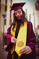 A person wearing a maroon graduation cap and gown with a bright yellow stole stands outdoors. The individual is holding the end of the stole, which features a university seal. The background includes modern buildings and tall cacti.
