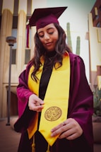 A person wearing a maroon graduation cap and gown with a bright yellow stole stands outdoors. The individual is holding the end of the stole, which features a university seal. The background includes modern buildings and tall cacti.