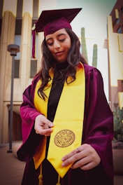 A person wearing a maroon graduation cap and gown with a bright yellow stole stands outdoors. The individual is holding the end of the stole, which features a university seal. The background includes modern buildings and tall cacti.