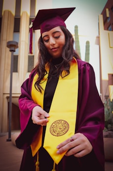 A person wearing a maroon graduation cap and gown with a bright yellow stole stands outdoors. The individual is holding the end of the stole, which features a university seal. The background includes modern buildings and tall cacti.