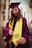 A person wearing a maroon graduation cap and gown with a bright yellow stole stands outdoors. The individual is holding the end of the stole, which features a university seal. The background includes modern buildings and tall cacti.