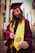 A person wearing a maroon graduation cap and gown with a bright yellow stole stands outdoors. The individual is holding the end of the stole, which features a university seal. The background includes modern buildings and tall cacti.