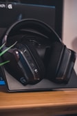 A pair of over-ear headphones rests on a tabletop stand. The headphones have a sleek, modern design with cushioned ear pads and buttons on the side. They are primarily black with some metallic accents. The backdrop appears to be part of a box with a logo visible.