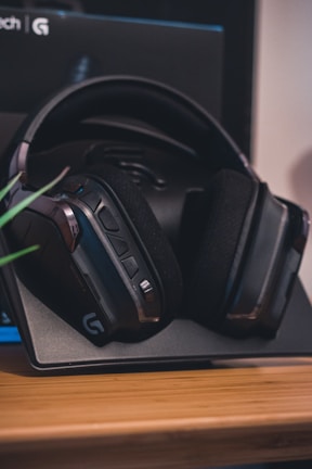 A pair of over-ear headphones rests on a tabletop stand. The headphones have a sleek, modern design with cushioned ear pads and buttons on the side. They are primarily black with some metallic accents. The backdrop appears to be part of a box with a logo visible.