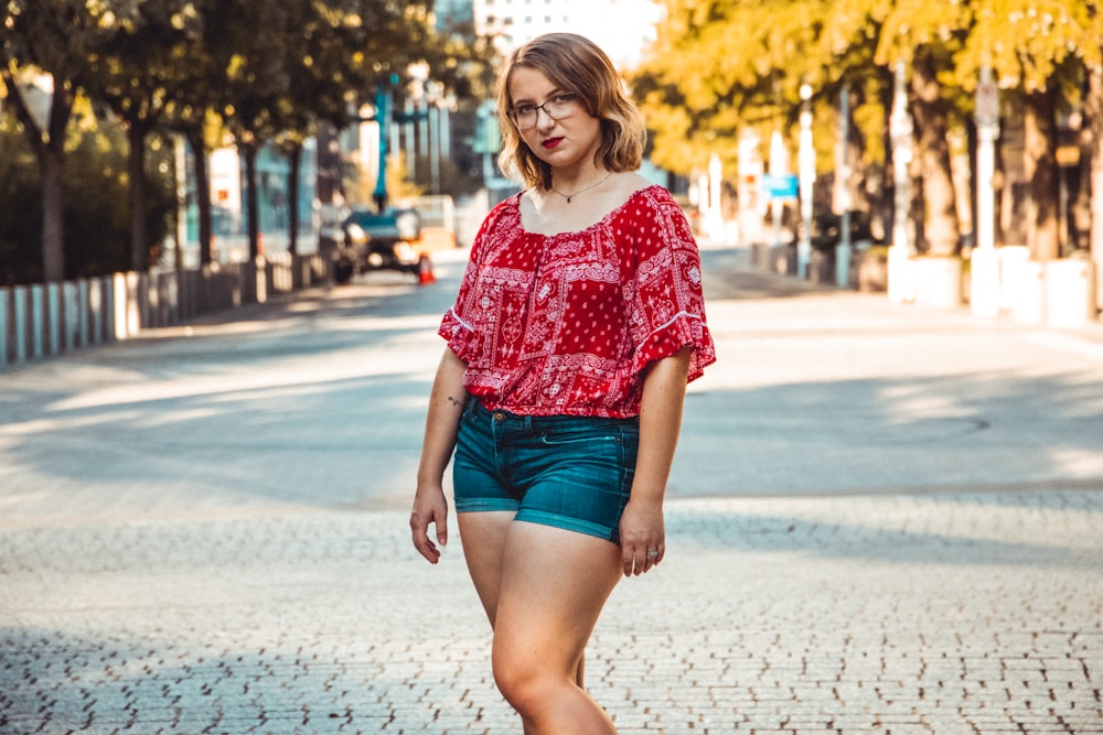 woman in red and white floral shirt and blue denim shorts standing on sidewalk during daytime