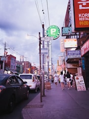 A busy urban street scene with various signage including a prominent Municipal Parking sign. People are walking on the sidewalk past shops and restaurants with colorful signage in different languages. Cars are parked along the street under a cloudy sky.