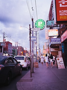 A busy urban street scene with various signage including a prominent Municipal Parking sign. People are walking on the sidewalk past shops and restaurants with colorful signage in different languages. Cars are parked along the street under a cloudy sky.