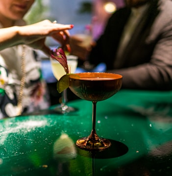 A dimly lit setting featuring a fancy glass cocktail garnished with a lime and a pink flower. Two individuals are interacting near the bar, with one person adjusting the garnish on the drink. The background is blurred, conveying a warm and inviting atmosphere.
