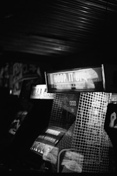 A black and white photograph of arcade machines with visible signage. The text 'Hoop It Up' is prominently displayed on one of the machines. The image captures the reflective surfaces and dimly lit arcade environment, with some graphics and scores visible on the machines.