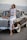 A woman in a white embroidered sleeveless top and white jeans leans against a vintage car. She is wearing sunglasses, hoop earrings, and open-toe block heels. A brown hat rests on the car's hood, and the background features a scenic landscape with hills.