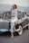 A woman in a white embroidered sleeveless top and white jeans leans against a vintage car. She is wearing sunglasses, hoop earrings, and open-toe block heels. A brown hat rests on the car's hood, and the background features a scenic landscape with hills.