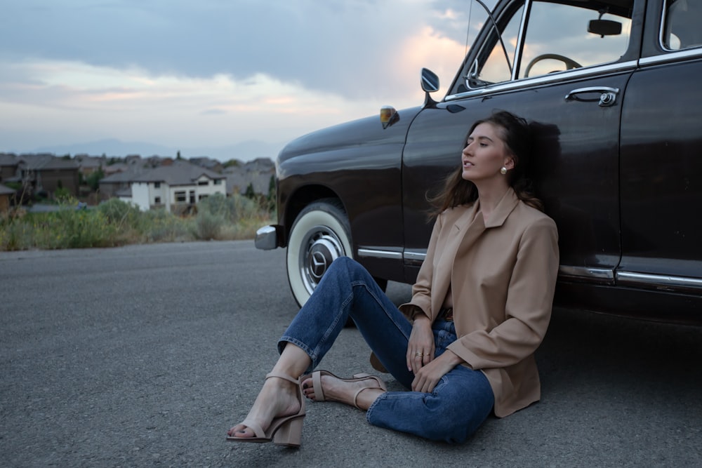 woman in brown coat and blue denim jeans sitting on black car hood during daytime