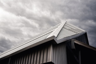 A modern building's roof with a sloped, triangular metal design is captured under a moody, overcast sky. The metallic surface of the roof contrasts with the dark, cloudy background, highlighting its architectural features.