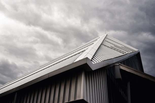 A modern building's roof with a sloped, triangular metal design is captured under a moody, overcast sky. The metallic surface of the roof contrasts with the dark, cloudy background, highlighting its architectural features.
