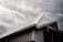 A modern building's roof with a sloped, triangular metal design is captured under a moody, overcast sky. The metallic surface of the roof contrasts with the dark, cloudy background, highlighting its architectural features.