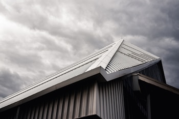 A modern building's roof with a sloped, triangular metal design is captured under a moody, overcast sky. The metallic surface of the roof contrasts with the dark, cloudy background, highlighting its architectural features.