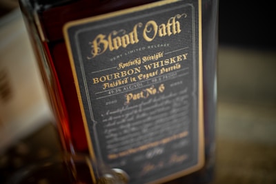 A close-up view of a bourbon whiskey bottle labeled 'Blood Oath'. The label is dark with elegant gold lettering detailing Kentucky straight bourbon whiskey finished in cognac barrels. It indicates details like 'very limited release' and '49.3% ALC/VOL'.