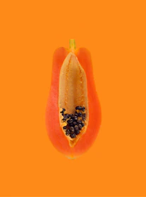 A halved papaya resting on a dark stone slab, its bright orange flesh and black seeds illuminated like a piece of fine art.