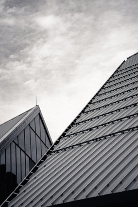 Modern architecture featuring triangular rooftops with metal cladding and a building façade composed of large glass panels under a cloudy sky.