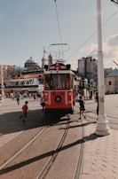 A red vintage tram marked 'Taksim Tünel' is moving along a tram track in a busy street. Several children are seen playfully interacting with the tram as it travels. In the background, there's a mix of old buildings, including a domed structure, and modern architecture. The sky is clear with a few clouds.