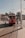 A red vintage tram marked 'Taksim Tünel' is moving along a tram track in a busy street. Several children are seen playfully interacting with the tram as it travels. In the background, there's a mix of old buildings, including a domed structure, and modern architecture. The sky is clear with a few clouds.
