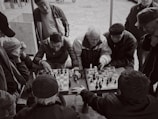 A group of men gathered around a small table, intently watching and playing chess. The scene is captured in black and white, giving it a timeless, nostalgic feel. The expressions on their faces show concentration and engagement, with one player making a move while others observe closely. The setting appears to be outdoors, possibly in a park or public space.