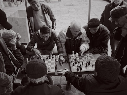A group of men gathered around a small table, intently watching and playing chess. The scene is captured in black and white, giving it a timeless, nostalgic feel. The expressions on their faces show concentration and engagement, with one player making a move while others observe closely. The setting appears to be outdoors, possibly in a park or public space.