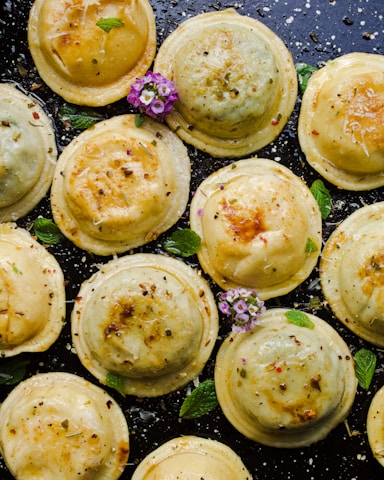 Several golden-brown ravioli pieces sprinkled with herbs and spices are arranged on a dark surface. Fresh mint leaves and a few small purple and white flowers provide contrast and garnish.