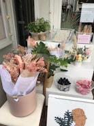 A display with a variety of plants and dried flowers is arranged on a set of white tables. There are bouquets wrapped in pastel-colored paper, a potted green plant, and two wire baskets containing small bottles and a pink fabric. A framed art print with a botanical theme sits at the front.