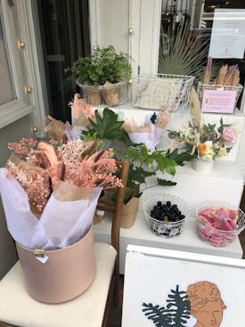 A display with a variety of plants and dried flowers is arranged on a set of white tables. There are bouquets wrapped in pastel-colored paper, a potted green plant, and two wire baskets containing small bottles and a pink fabric. A framed art print with a botanical theme sits at the front.