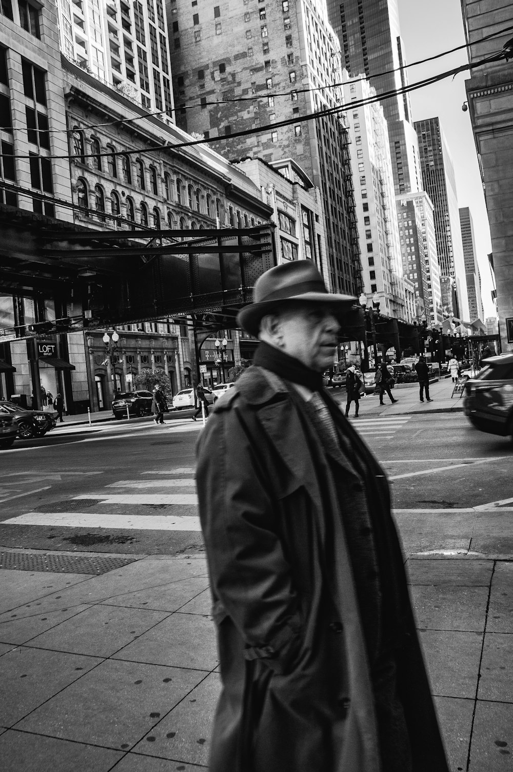 grayscale photo of man in black coat and hat standing on sidewalk
