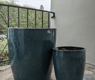 Two large, glossy ceramic planters, one taller than the other, are placed on a tiled floor next to a metal railing. The background shows a lush green hedge and part of a white wall.