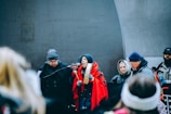 A group of people dressed warmly, some wearing hats and gloves, are gathered around a microphone. One individual wrapped in a red shawl appears to be speaking passionately, holding a traditional drum. Others in the background seem attentive and focused.
