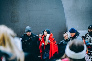 A group of people dressed warmly, some wearing hats and gloves, are gathered around a microphone. One individual wrapped in a red shawl appears to be speaking passionately, holding a traditional drum. Others in the background seem attentive and focused.