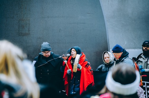 A group of people dressed warmly, some wearing hats and gloves, are gathered around a microphone. One individual wrapped in a red shawl appears to be speaking passionately, holding a traditional drum. Others in the background seem attentive and focused.