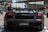 A sleek black Lamborghini sports car is parked outdoors. The focus is on the rear of the car, showcasing its aerodynamic design and distinctive taillights. There are people walking in the background, and modern architectural structures are visible.