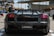 A sleek black Lamborghini sports car is parked outdoors. The focus is on the rear of the car, showcasing its aerodynamic design and distinctive taillights. There are people walking in the background, and modern architectural structures are visible.