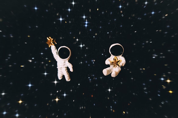 Two small toy astronauts float in a dark, starry space. One holds a golden star in an outstretched hand, while the other cradles a star in both hands. The background is filled with numerous bright, scattered stars on a black backdrop.