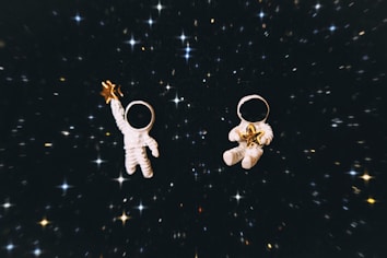 Two small toy astronauts float in a dark, starry space. One holds a golden star in an outstretched hand, while the other cradles a star in both hands. The background is filled with numerous bright, scattered stars on a black backdrop.