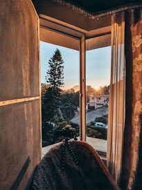 A cozy interior scene featuring an open window with sheer curtains, through which a tranquil sunset view is visible. A pine tree is prominently framed against the sky, accompanied by distant houses and a road. A camera on a tripod is set up indoors, aimed at the scene outside. The warm glow of the sunset bathes the room, creating a serene atmosphere.