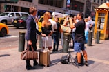 Two people are standing on a city sidewalk with luggage, engaged in conversation. A man nearby is preparing to take a photo, setting up a tripod and camera. Several others are visible in the background, along with yellow cabs and parked cars. The scene takes place in a busy urban area during the day.