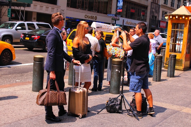 Two people are standing on a city sidewalk with luggage, engaged in conversation. A man nearby is preparing to take a photo, setting up a tripod and camera. Several others are visible in the background, along with yellow cabs and parked cars. The scene takes place in a busy urban area during the day.