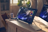 A bright studio space features a drawing tablet on a wooden desk displaying digital art in progress. The image on the screen shows a character with long blonde hair and intricate clothing. Natural light streams through a window illuminating the workspace. Various art tools like a color palette and rolled papers are scattered around, alongside a computer monitor.