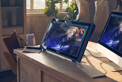 A bright studio space features a drawing tablet on a wooden desk displaying digital art in progress. The image on the screen shows a character with long blonde hair and intricate clothing. Natural light streams through a window illuminating the workspace. Various art tools like a color palette and rolled papers are scattered around, alongside a computer monitor.