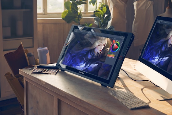A bright studio space features a drawing tablet on a wooden desk displaying digital art in progress. The image on the screen shows a character with long blonde hair and intricate clothing. Natural light streams through a window illuminating the workspace. Various art tools like a color palette and rolled papers are scattered around, alongside a computer monitor.