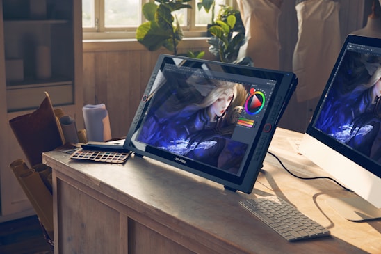A bright studio space features a drawing tablet on a wooden desk displaying digital art in progress. The image on the screen shows a character with long blonde hair and intricate clothing. Natural light streams through a window illuminating the workspace. Various art tools like a color palette and rolled papers are scattered around, alongside a computer monitor.