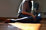 A person in athletic wear is sitting on a polished wooden floor with their legs crossed. Warm light filters through a window, casting a soft glow. A mirrored wall reflects part of the figure and some additional room features.