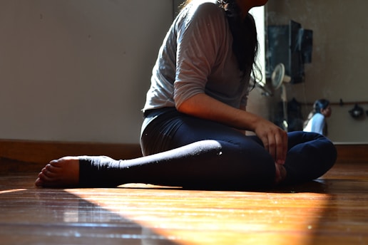 A person in athletic wear is sitting on a polished wooden floor with their legs crossed. Warm light filters through a window, casting a soft glow. A mirrored wall reflects part of the figure and some additional room features.