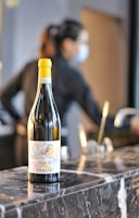 A wine bottle with a yellow cap is placed on a marble counter. The label on the bottle is detailed and colorful, suggesting it is a Moscato d’Asti. In the blurred background, a person wearing a black outfit and a face mask can be seen, suggesting an indoor setting, possibly a bar or restaurant.